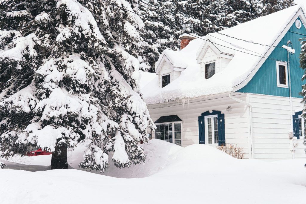Ett blått trähus med djup snö på tomten och snötyngda granar